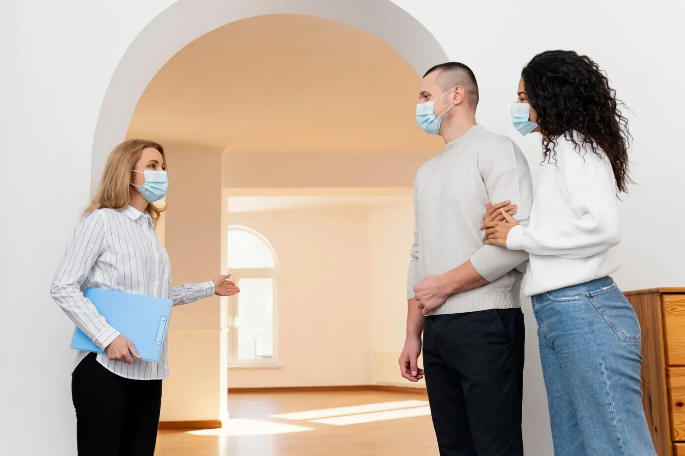 A female realtor wearing a medical mask is showing a couple a new house.